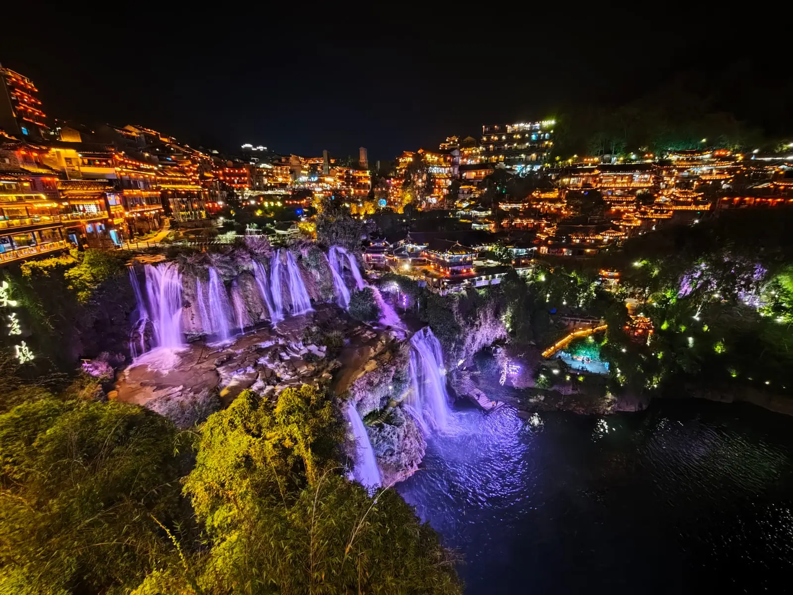 El pueblo antiguo de Furong iluminado de noche, con su cascada y las casas de madera colgadas sobre el agua